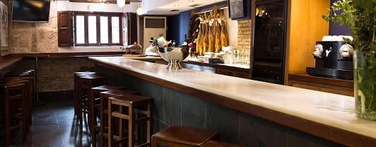 El interior de La Taberna de Marisa: una barra larga con taburetes de madera, paredes de piedra y jamones colgados detrás de la barra.